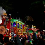 Crowd reaching for throws as a lit-up steam train float rolls through a nighttime Mardi Gras parade in New Orleans