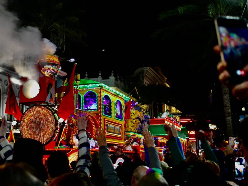 Crowd reaching for throws as a lit-up steam train float rolls through a nighttime Mardi Gras parade in New Orleans
